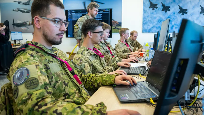 Zes militairen kijken aan een bureau geconcentreerd naar enkele computerschermen. 