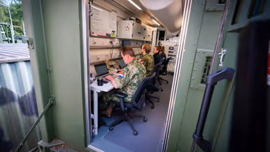 Vier personen, van wie twee in militair uniform, zitten achter beeldschermen in een groene container.