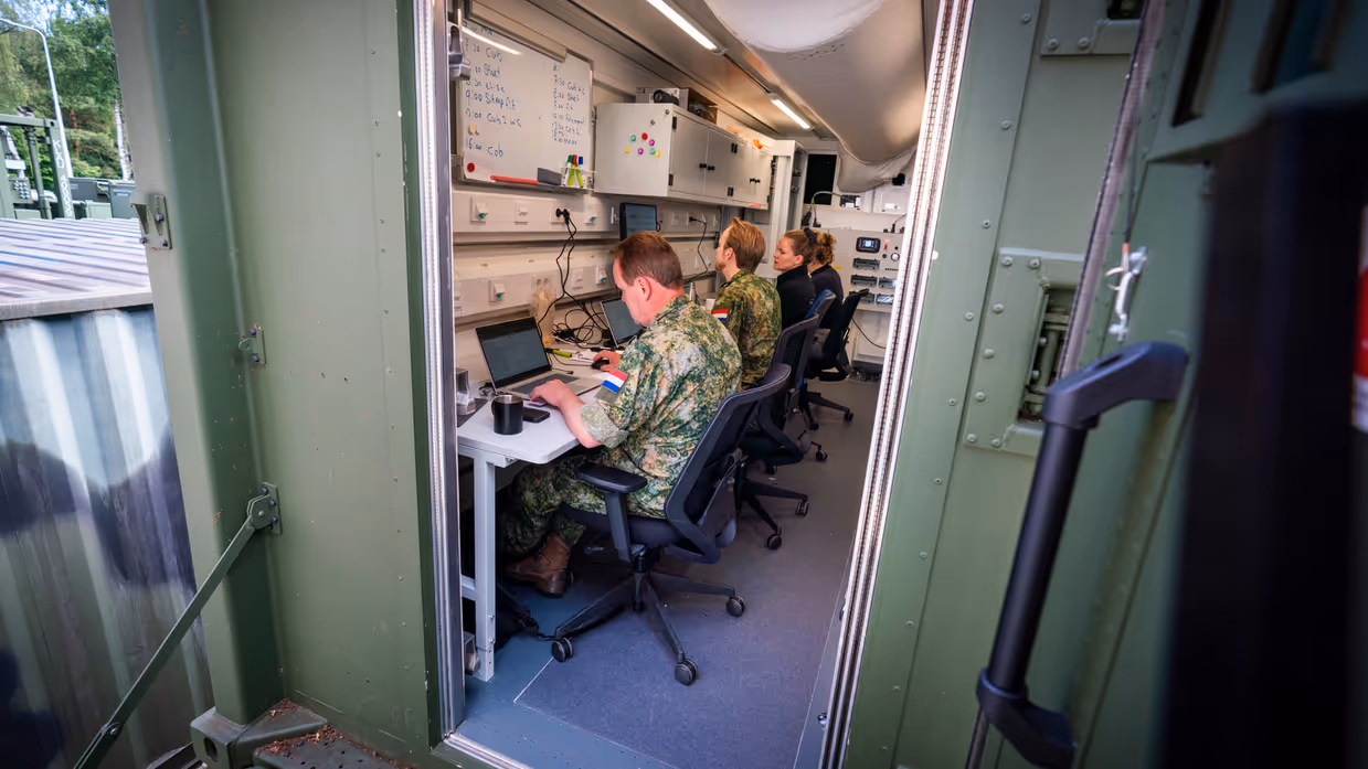 Vier personen, van wie twee in militair uniform, zitten achter beeldschermen in een groene container.