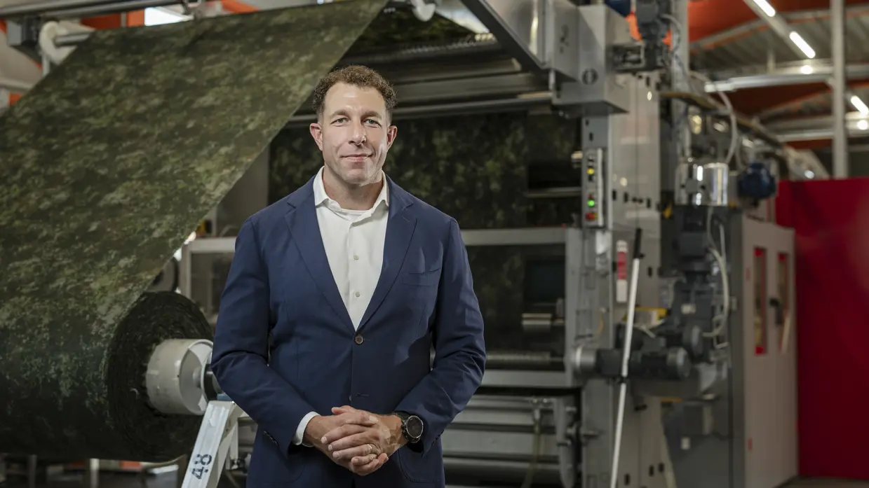 Een man met blauwe colbertjas poseert in een fabriekshal. Op de achtergrond wordt een grote rol stof in camouflagepatroon door een machine geleid.