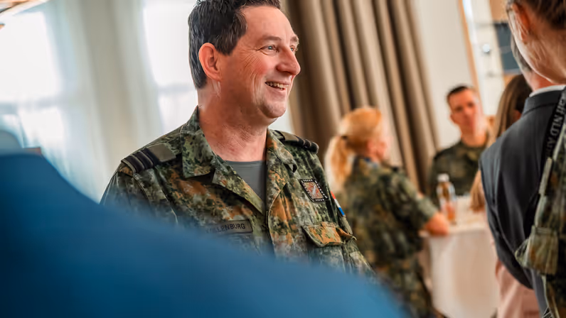 Een lachende militair met op de achtergrond 2 collega’s aan een tafel. 