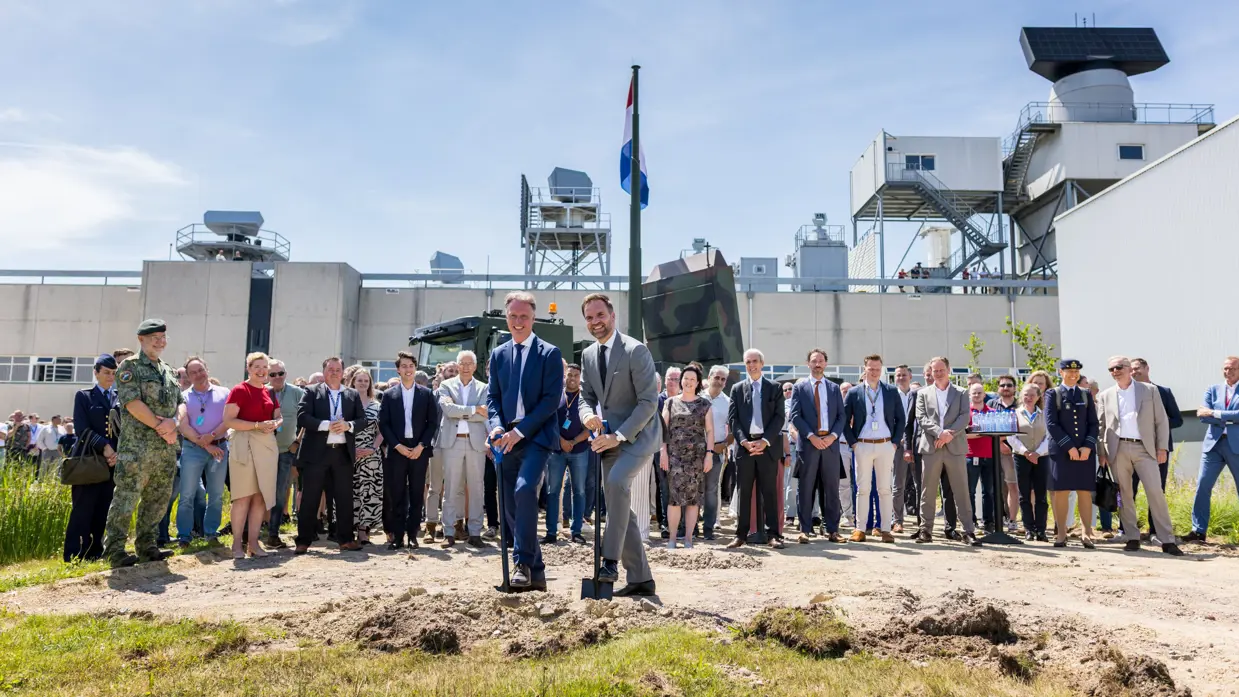  Staatssecretaris Derk Boswijk kijkt in de camera terwijl hij een schop in de grond zet. Achter hem een groep mensen die toekijkt.
