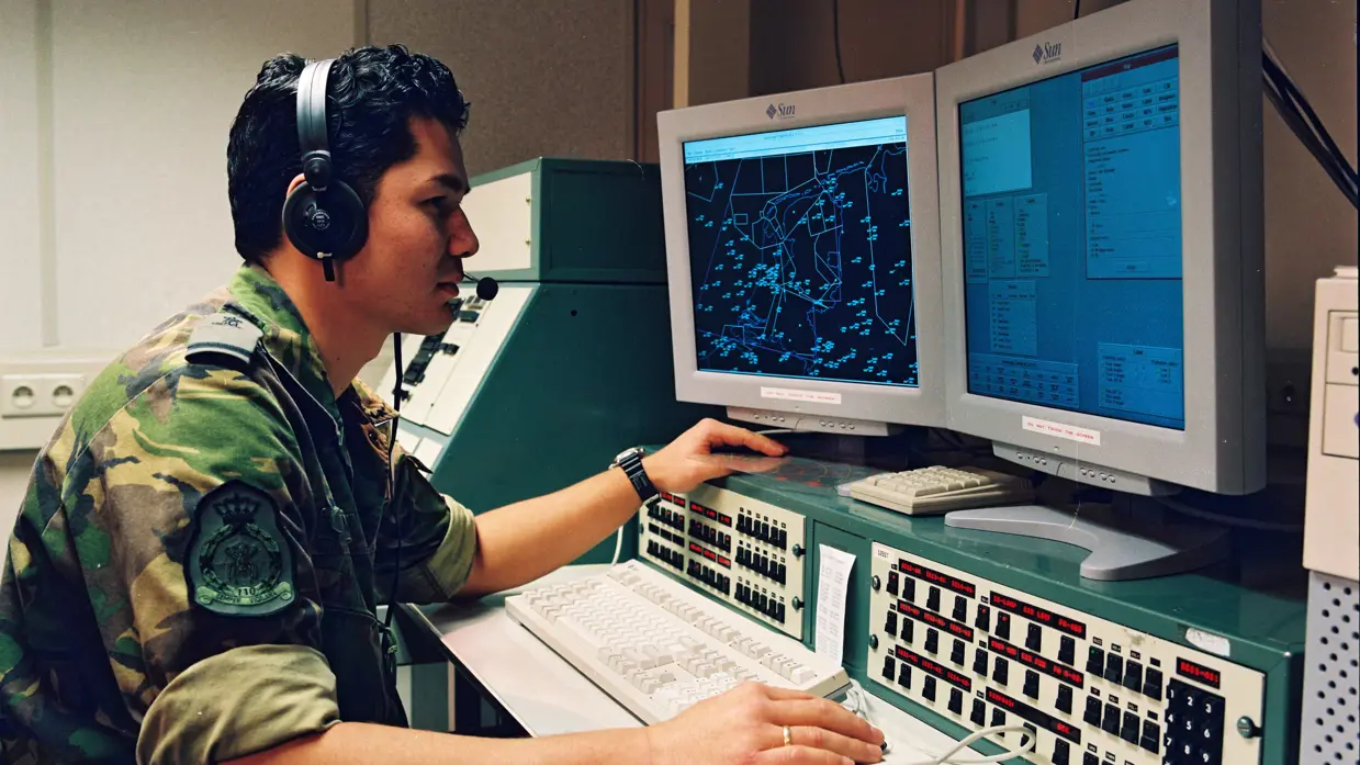 Een militair in camouflagepak achter twee computerschermen. 