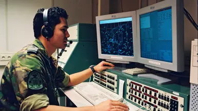 Een militair in camouflagepak achter twee computerschermen. 