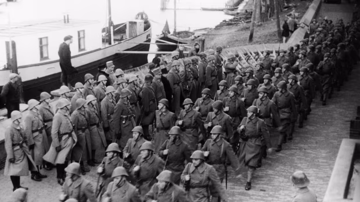 Een groep soldaten in uniform met helmen, marcherend langs een kade met een boot en omstanders op de achtergrond. De foto is in zwart-wit.