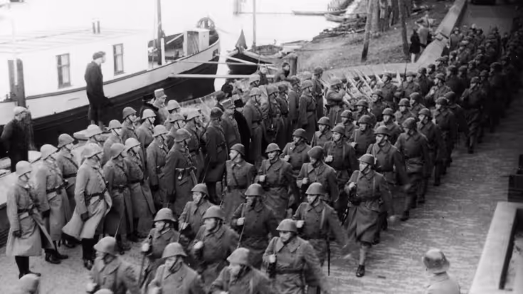 Een groep soldaten in uniform met helmen, marcherend langs een kade met een boot en omstanders op de achtergrond. De foto is in zwart-wit.