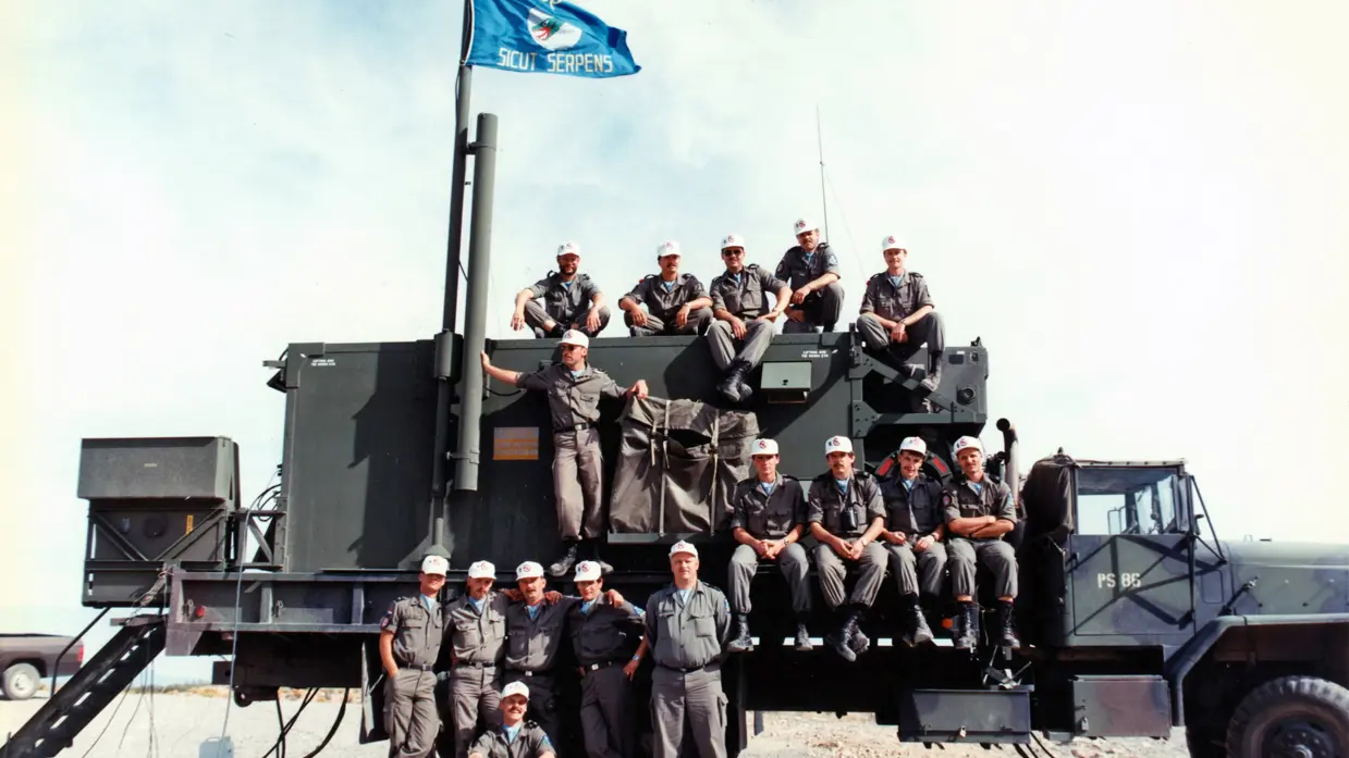 Een groep militairen zit op een groene vrachtwagen. Aan de mast wappert een squadron-vlag