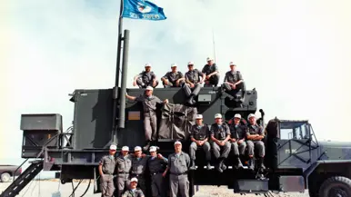 Een groep militairen zit op een groene vrachtwagen. Aan de mast wappert een squadron-vlag