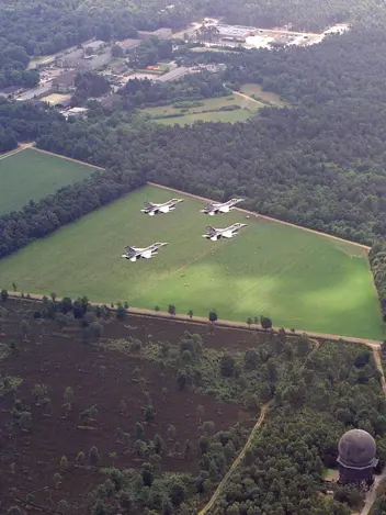Vier F-16’s vliegen in formatie boven een bos- en heidegebied. Rechtsonder is een radarbol te zien. Linksboven staan gebouwen.
