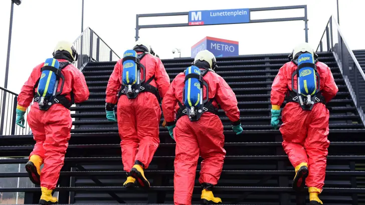 Personen in rode beschermende pakken en gasflessen op de rug lopen trap van een metrostation op.