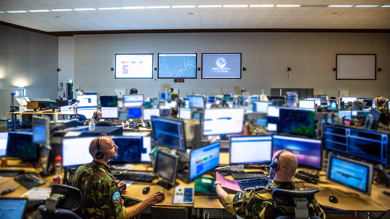  Overzichtsfoto van een ruimte met veel computerschermen. In de achtergrond op de muur hangen drie grote projectieschermen. Foto: 