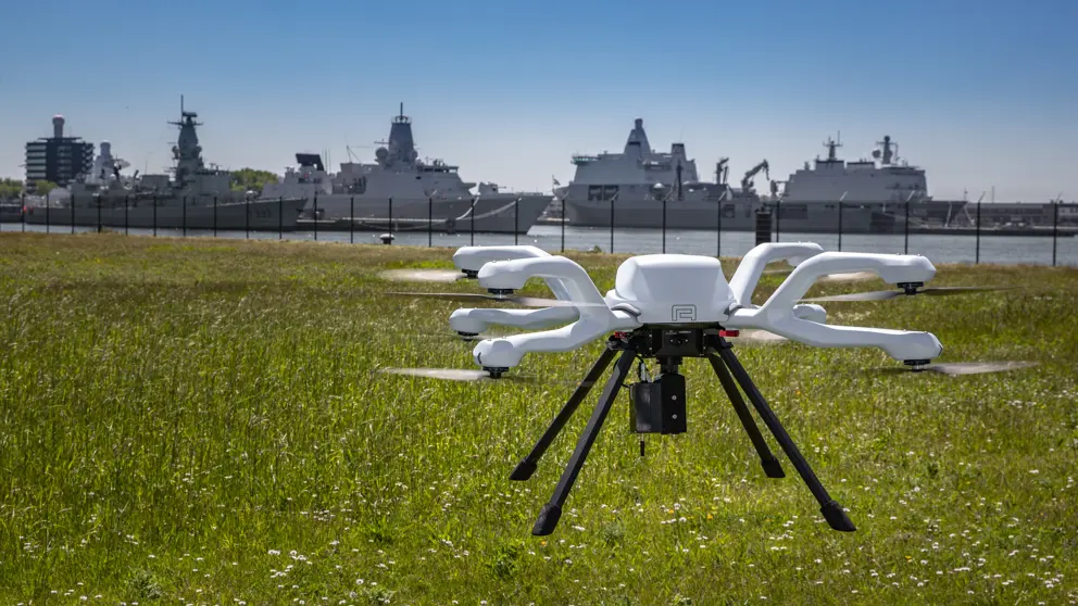 Een witte drone vliegt boven een grasveld, met op de achtergrond meerdere marineschepen aangemeerd in een haven.
