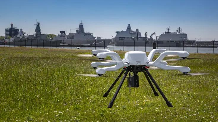 Een witte drone vliegt boven een grasveld, met op de achtergrond meerdere marineschepen aangemeerd in een haven.