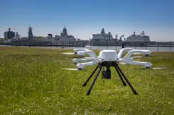 Een witte drone vliegt boven een grasveld, met op de achtergrond meerdere marineschepen aangemeerd in een haven.