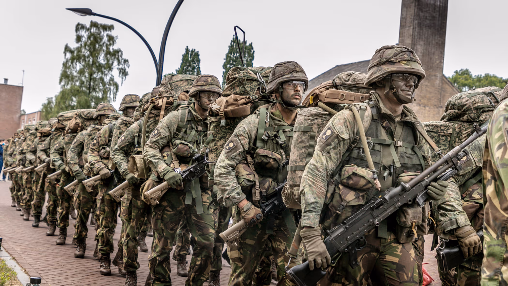 Militairen marcheren langs de fotograaf. Ze dragen een wapen, volle bepakking en hebben camouflage op het gezicht. 