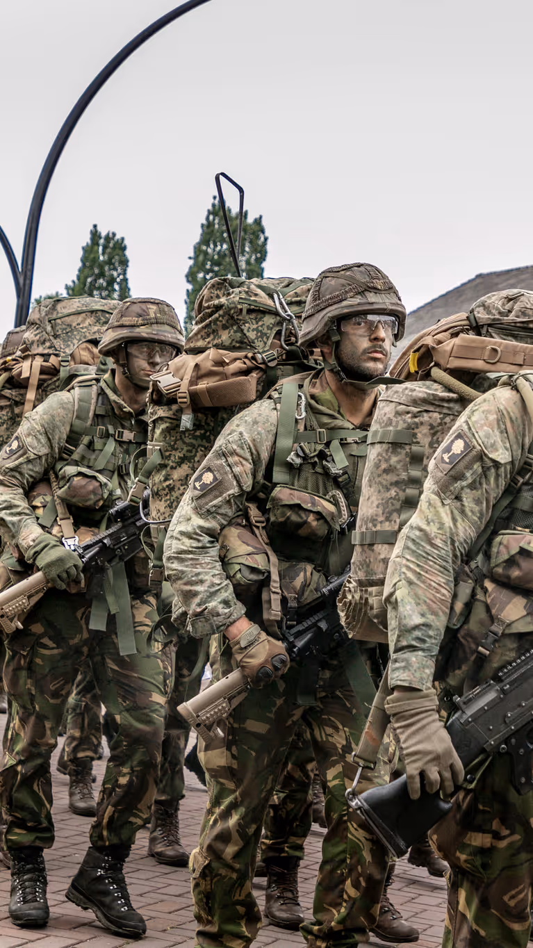 Militairen marcheren langs de fotograaf. Ze dragen een wapen, volle bepakking en hebben camouflage op het gezicht. 