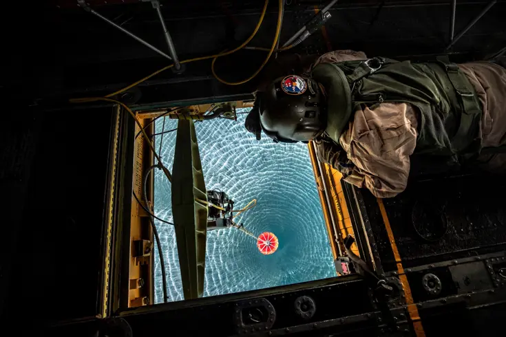 Een loadmaster hangt boven een luik in een helikopter en checkt de bambi bucket die water schept.