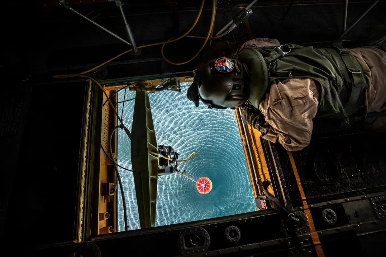 Een loadmaster hangt boven een luik in een helikopter en checkt de bambi bucket die water schept.