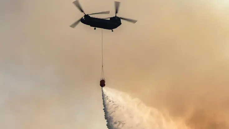 Chinook tegen oranje lucht stort water via waterzak onder het toestel.