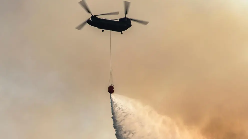 Chinook-transporthelikopter in oranjegekleurde lucht stort water uit met bambi-bucket onder het toestel.