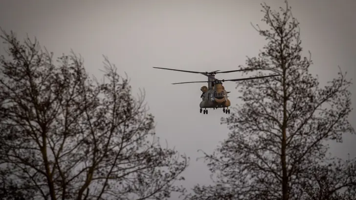 Een Chinook-transporthelikopter komt aangevlogen tussen 2 bomen.