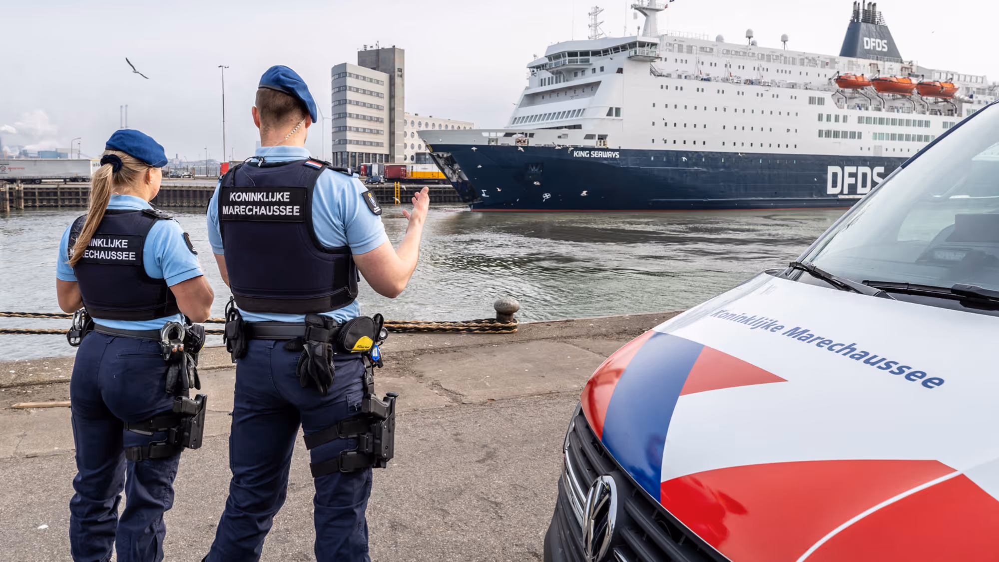 Marechausseevoertuig met 2 marechaussees ervoor. Zij kijken naar een schip in de haven.