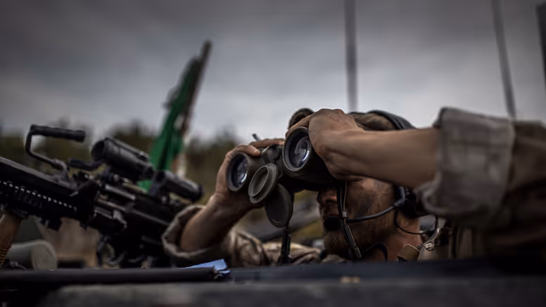 Een militair kijkt door een verrekijker. Hij draagt camouflage. Achter hem staat een wapen, een Colt C7.  