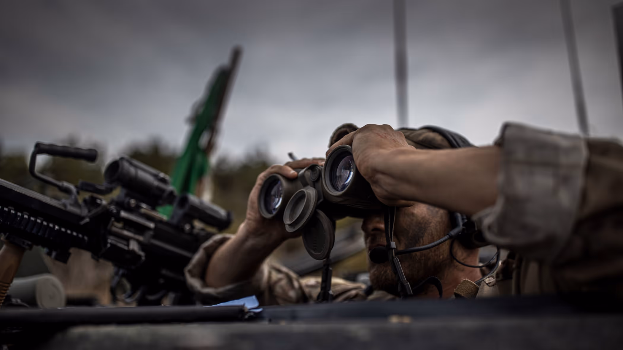 Een militair kijkt door een verrekijker. Hij draagt camouflage. Achter hem staat een wapen, een Colt C7.  
