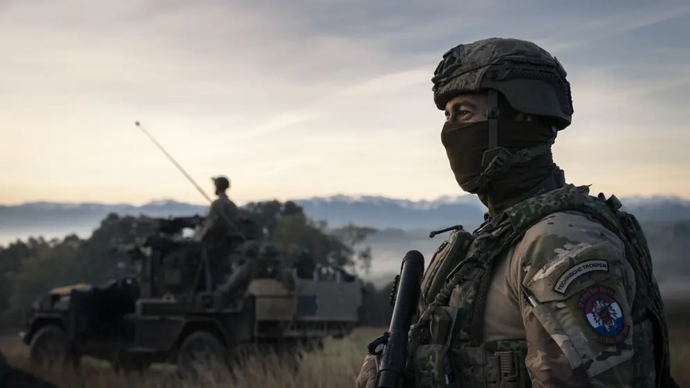 Militair met gezichtsbedekking op de uitkijk naast militair voertuig in natuurgebied.