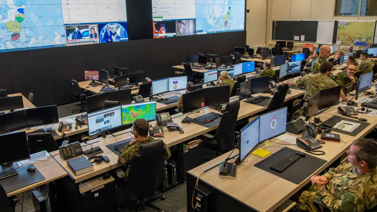 Een ruimte met in de achtergrond een meer dan levensgroot beeldscherm waarop diverse informatie wordt weergegeven. In de voorgrond zitten militairen uit diverse landen te werken achter computerschermen.