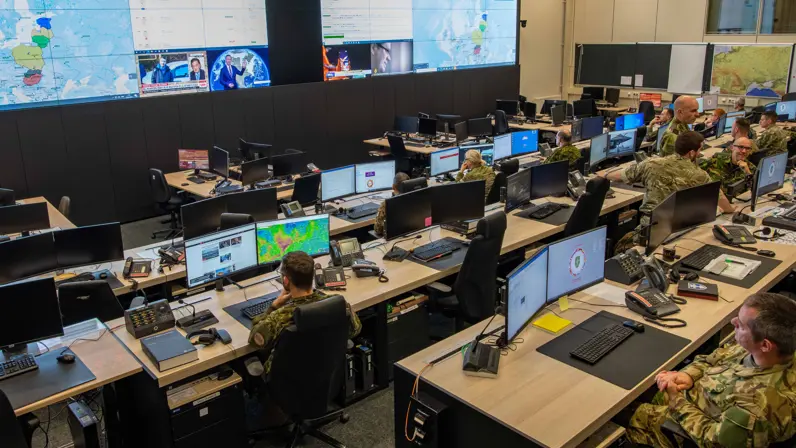Een ruimte met in de achtergrond een meer dan levensgroot beeldscherm waarop diverse informatie wordt weergegeven. In de voorgrond zitten militairen uit diverse landen te werken achter computerschermen.