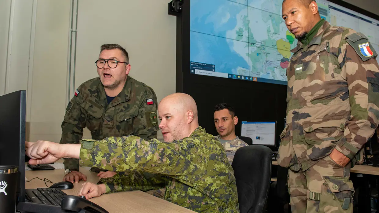 Een militair zit achter een computer. Drie militairen uit andere landen kijken over zijn schouder mee.