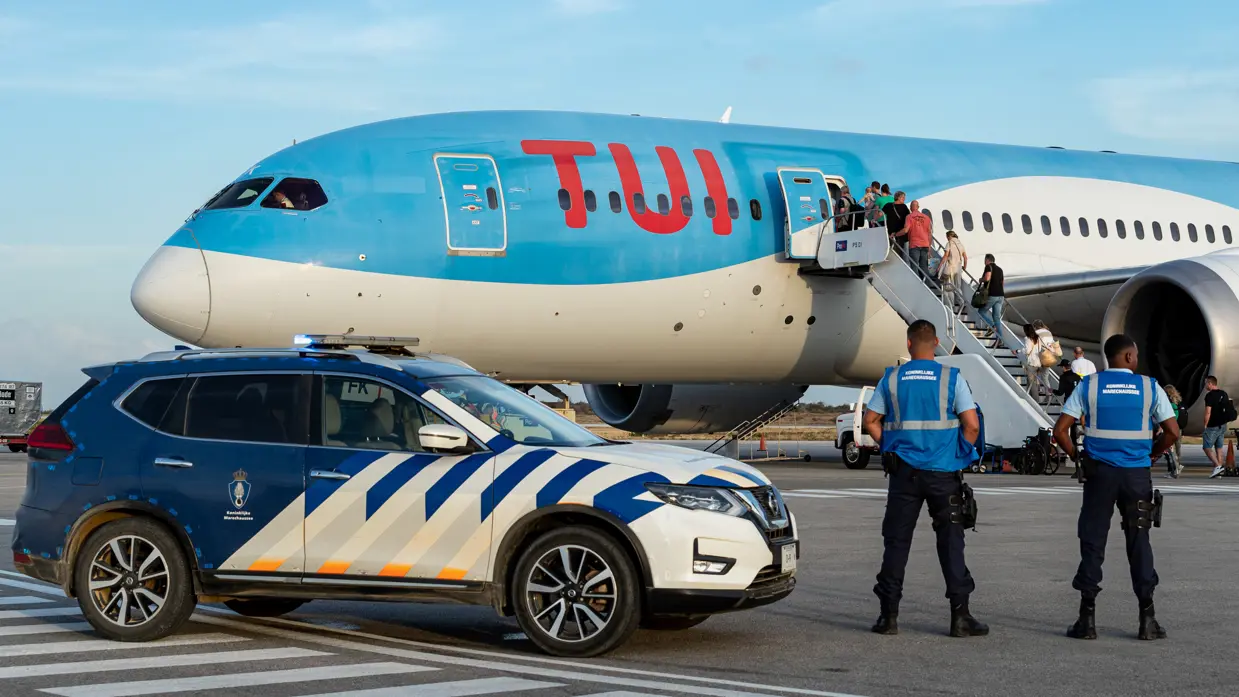 Marechaussees naast een dienstwagen op het vliegveld op Bonaire met een vliegtuig van TUI op de achtergrond waar passagiers instappen.