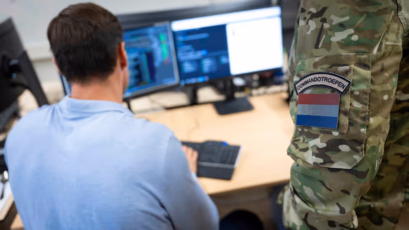 Een man zittend achter zijn computer, daarnaast een staande militair met op zijn arm de tekst ‘Korps Commandotroepen’.