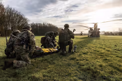 5 militairen zitten op hun knieën in het gras bij een gewonde op een brancard. 2 Chinook-transporthelikopters zijn zichtbaar op de achtergrond.