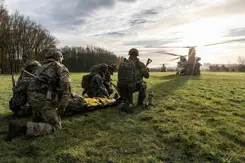 Militairen oefenen op een grasveld. 1 ligt op een brancard. Op de achtergrond 2 Chinook-transporthelikopters.