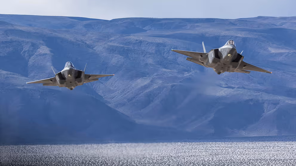2 F-35 jachtvliegtuigen vliegen boven water tegen een achtergrond van bergen in blauw licht.