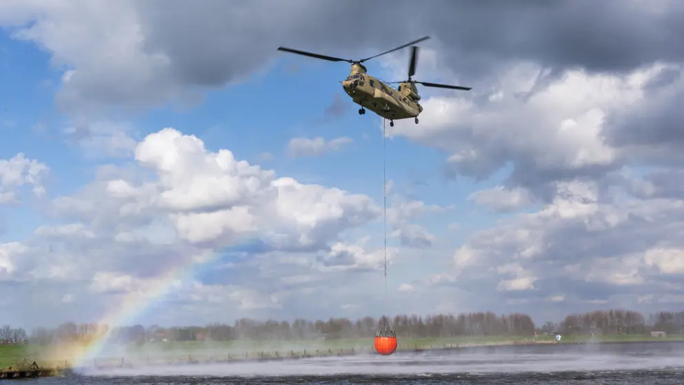 Chinook-transporthelikopter met rode bambi-bucket boven water tegen een blauwe lucht met witte wolken. Links is een regenboog te zien.