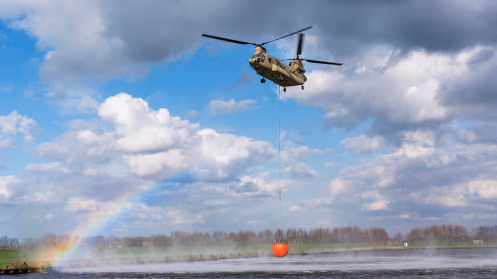 Chinook-transporthelikopter met rode bambi-bucket boven water tegen een blauwe lucht met witte wolken. Links is een regenboog te zien.