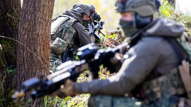 Een militair ligt achter zijn wapen en kijkt over zijn schouder. Achter hem zit een gewapende collega, die onscherp te zien is. 