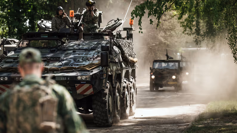 Een Boxer rijdt over een zanderig paadje, erachter rijdt een MB. Het zand stuift op. OP de voorgrond is de rug van een militair te zien.&nbsp;