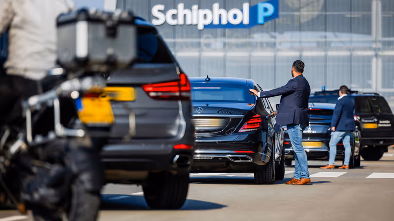 Een man met donkerblauw colbert en spijkerbroek opent het portier van een stilstaande auto. Voor en achter de auto staan eveneens voertuigen geparkeerd. Op de achtergrond is vliegveld Schiphol te zien.