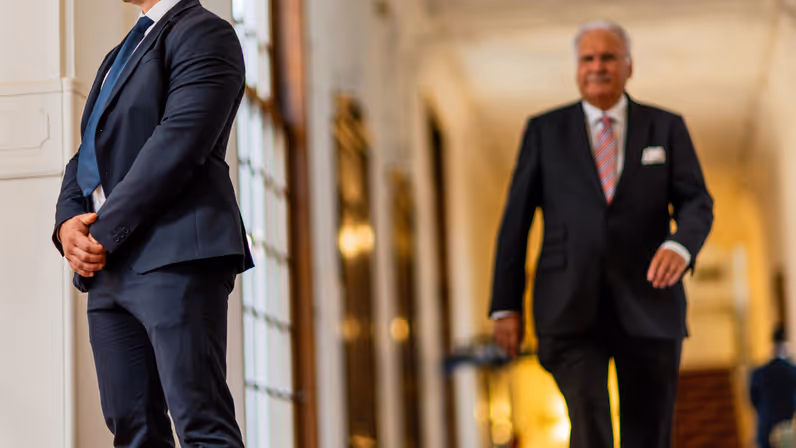 Een persoonsbeveiliger in donkerblauw pak kijkt voor zich uit, terwijl achter hem een oudere man met grijze snor aan komt lopen.