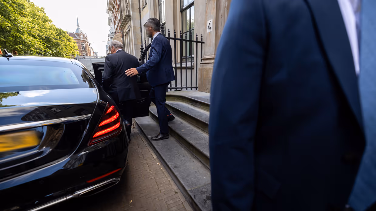 en man in donkerblauw pak begeleidt een oudere man een auto in. Dit voertuig heeft geblindeerde ramen.