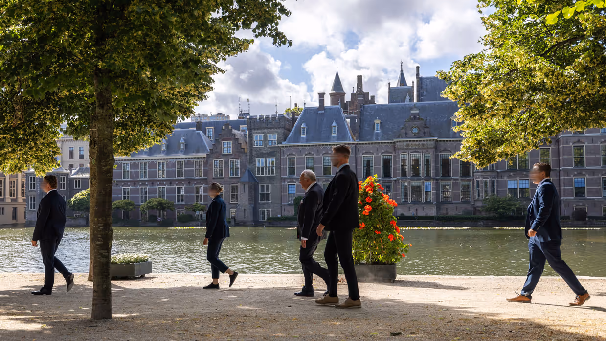 Drie persoonsbeveiligers staan in een gang en omringen een bellende man in pak. Daarnaast vier persoonsbeveiligers die rondom een man lopen bij het Binnenhof in Den Haag.