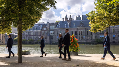 Drie persoonsbeveiligers staan in een gang en omringen een bellende man in pak. Daarnaast vier persoonsbeveiligers die rondom een man lopen bij het Binnenhof in Den Haag.