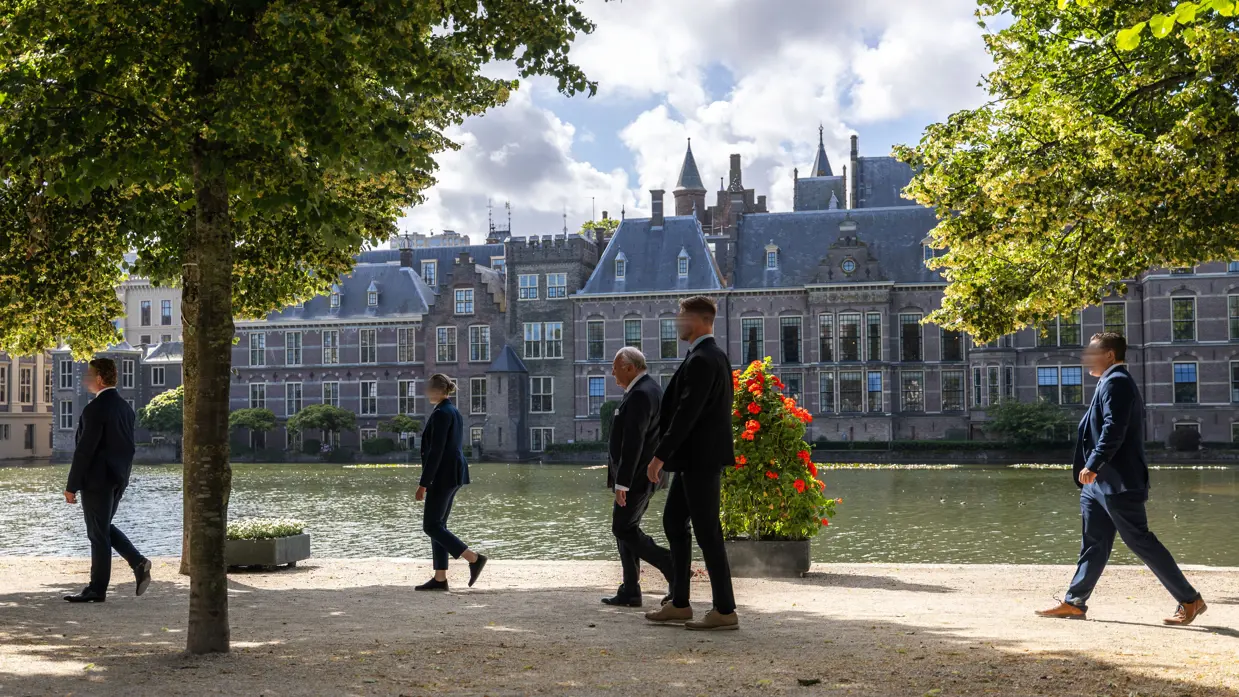 Drie persoonsbeveiligers staan in een gang en omringen een bellende man in pak. Daarnaast vier persoonsbeveiligers die rondom een man lopen bij het Binnenhof in Den Haag.
