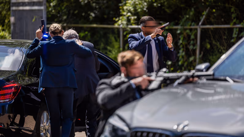 Drie persoonsbeveiligers achter een auto. Een van hen begeleidt de TBP de auto in, haar pistool in de lucht gericht. De twee daarvoor richten de andere kant op. De een met een pistool, de ander met een lang wapen.