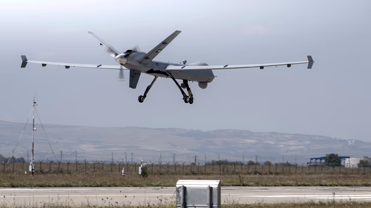 Een grijze MQ-9 Reaper, net boven een landingsbaan. Op de achtergrond zijn bergen te zien.