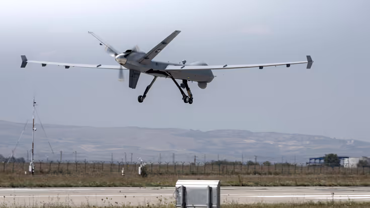 Een grijze MQ-9 Reaper, net boven een landingsbaan. Op de achtergrond zijn bergen te zien.